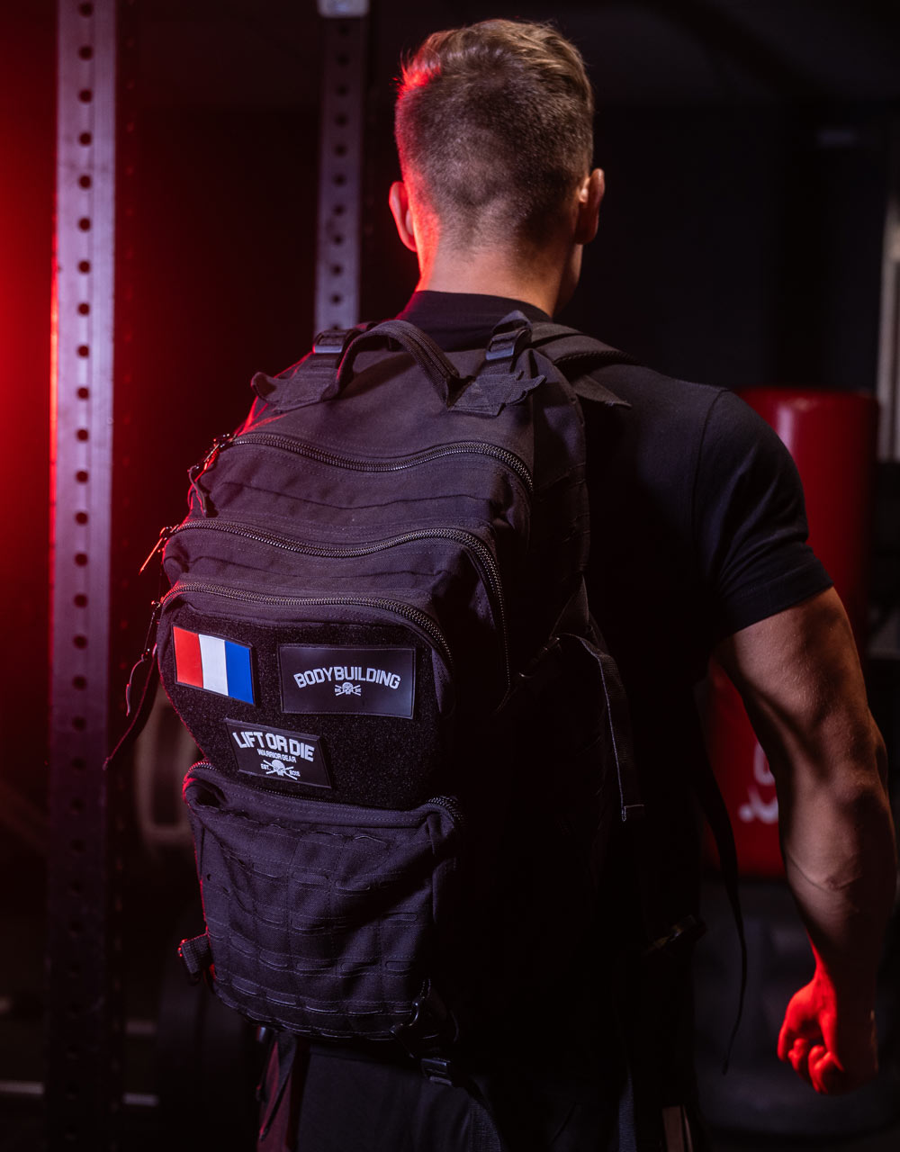 Athlet von hinten, der den schwarzen taktischen Bodybuilding-Rucksack Warrior Gear mit Klettpatches France, Bodybuilding und Lift or Dieträgt.